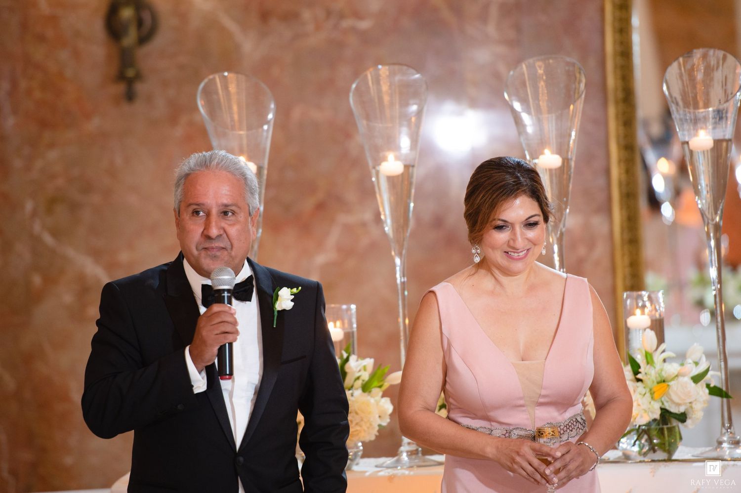 Elegant couple at wedding reception, man in tuxedo speaking into microphone, woman in pink gown standing beside tall glass candle holders.