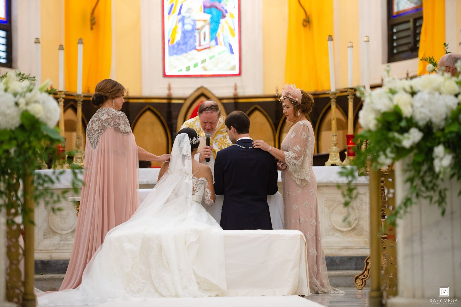 Bride and groom kneel at church altar surrounded by bridesmaids in pink gowns during wedding blessing ceremony.