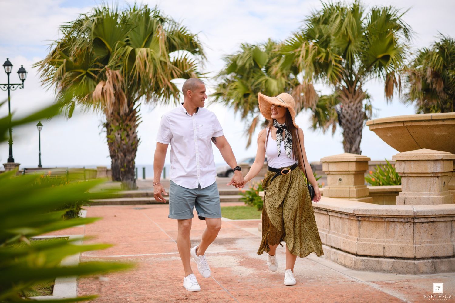 Couple holding hands walking along a tropical waterfront promenade with palm trees on a sunny day.