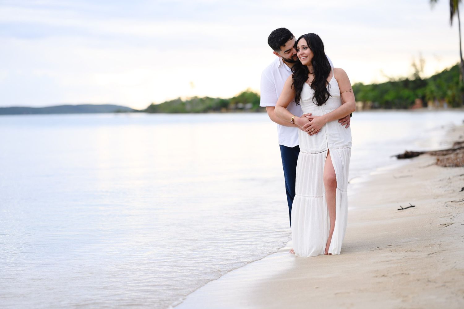 Couple embracing on a tropical beach at sunset, woman in white dress, man in navy pants, with calm ocean waters behind them.