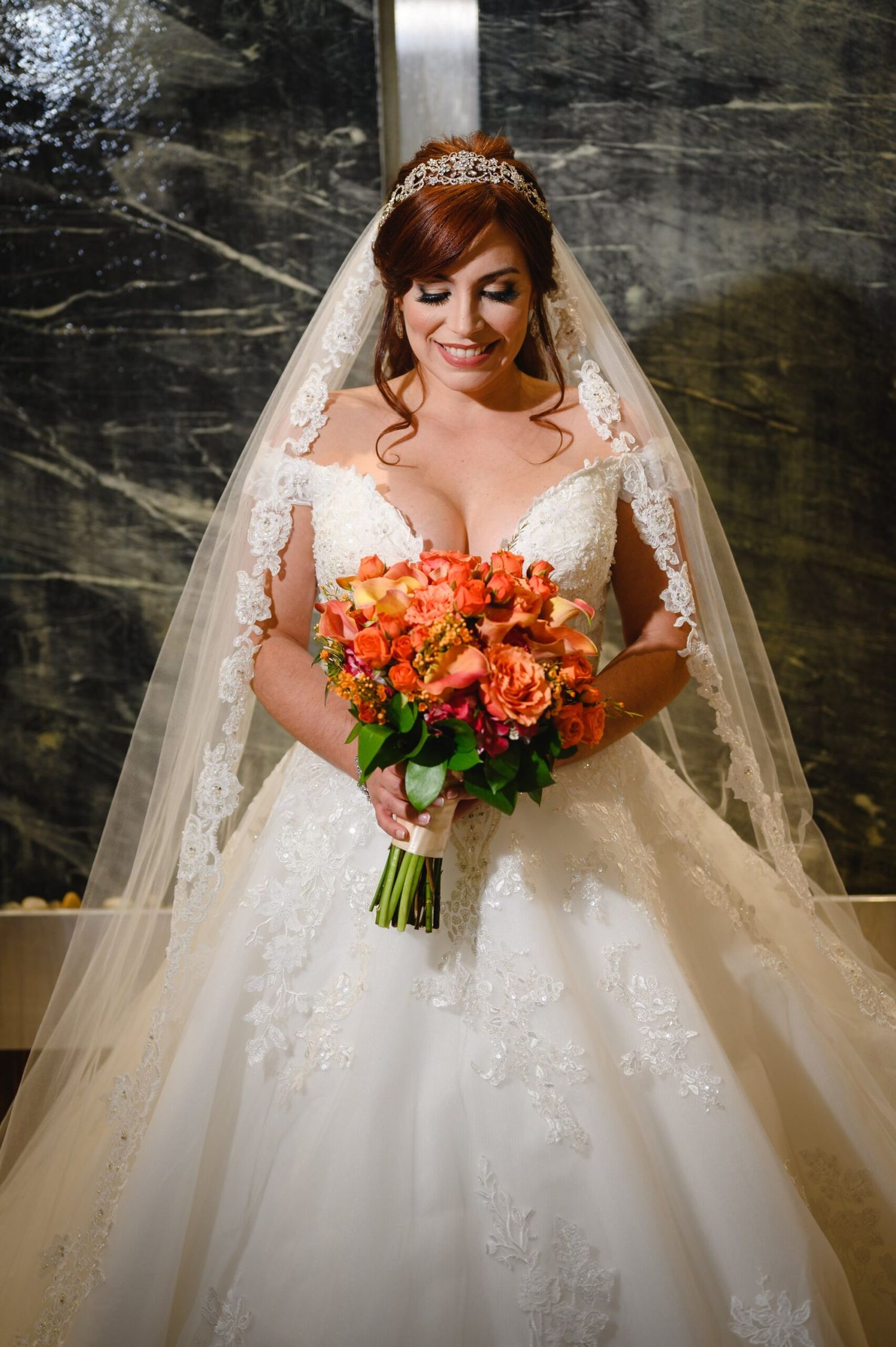 Bride in lace ball gown and veil holding orange rose bouquet, smiling downward in elegant marble venue.