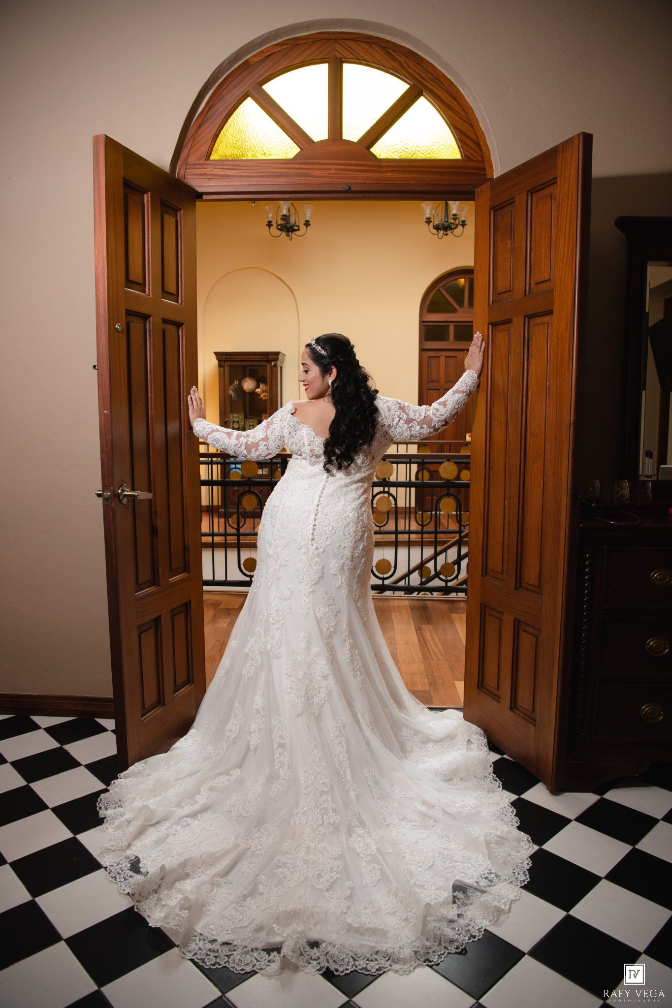 Bride in lace mermaid wedding gown with long train poses in open wooden double doors on black and white checkered floor.