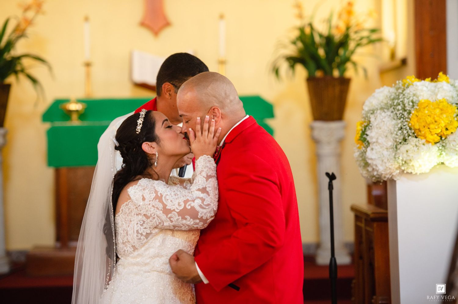 Bride and groom share first kiss at church altar, groom in red suit, bride in lace wedding dress.
