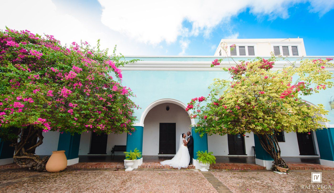 Bride and groom pose in front of a blue colonial building adorned with vibrant bougainvillea flowers.