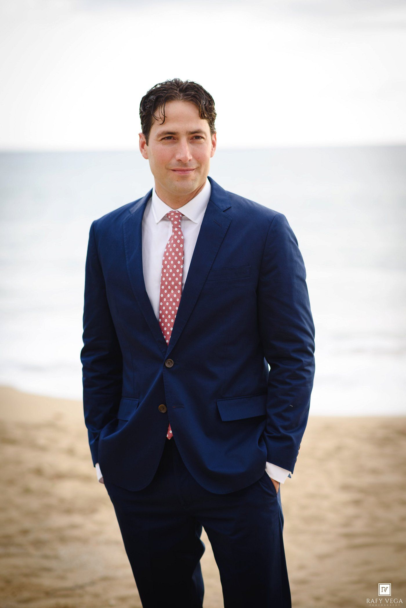 Man in navy blue suit and red polka dot tie posing on sandy beach with ocean backdrop.