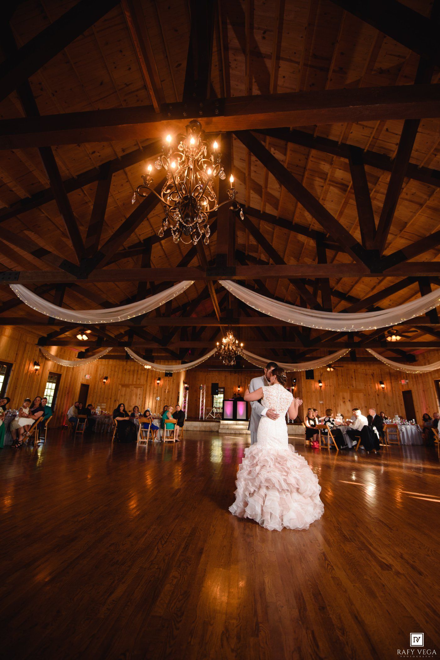 Bride and groom share first dance in rustic barn wedding venue with chandelier and draped fabric ceiling decorations.