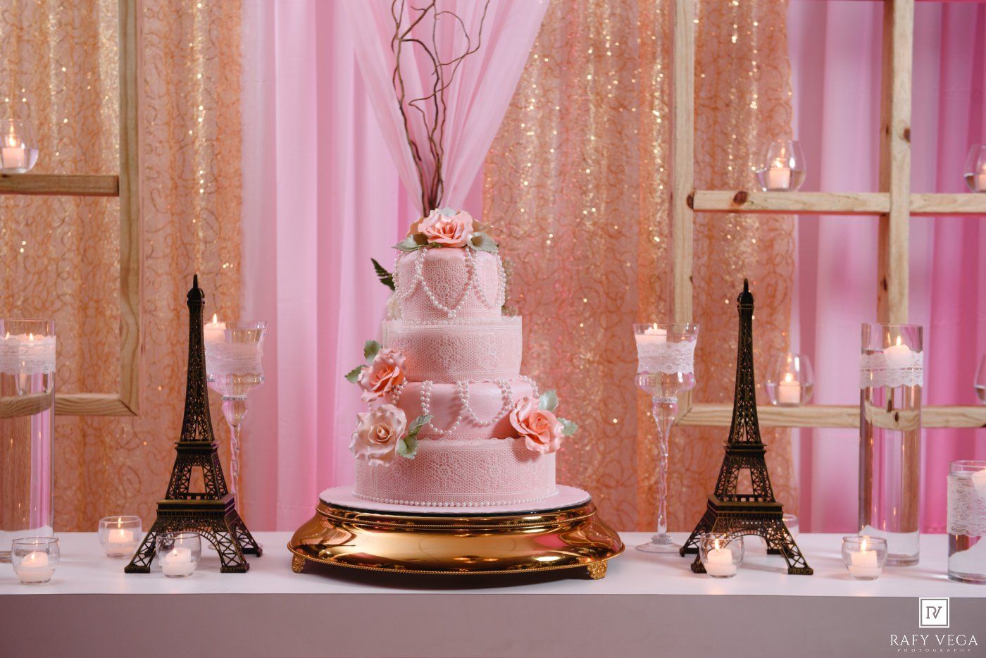 Elegant four-tier white wedding cake with pink roses flanked by Eiffel Tower figurines on a gold stand against a pink sequin backdrop.