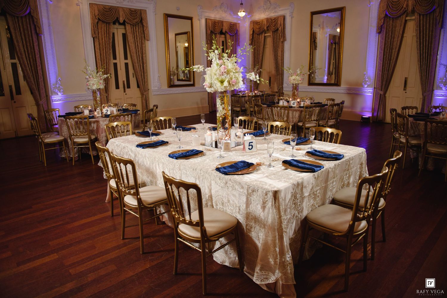 Elegant wedding reception hall with gold Chiavari chairs, ivory lace tablecloths, navy blue napkins, and tall floral centerpieces.