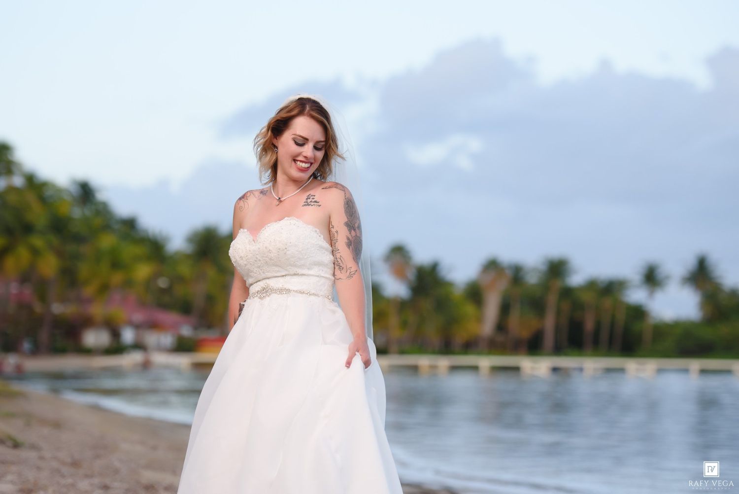 Bride in strapless white wedding gown smiling on tropical beach with palm trees and ocean in background.