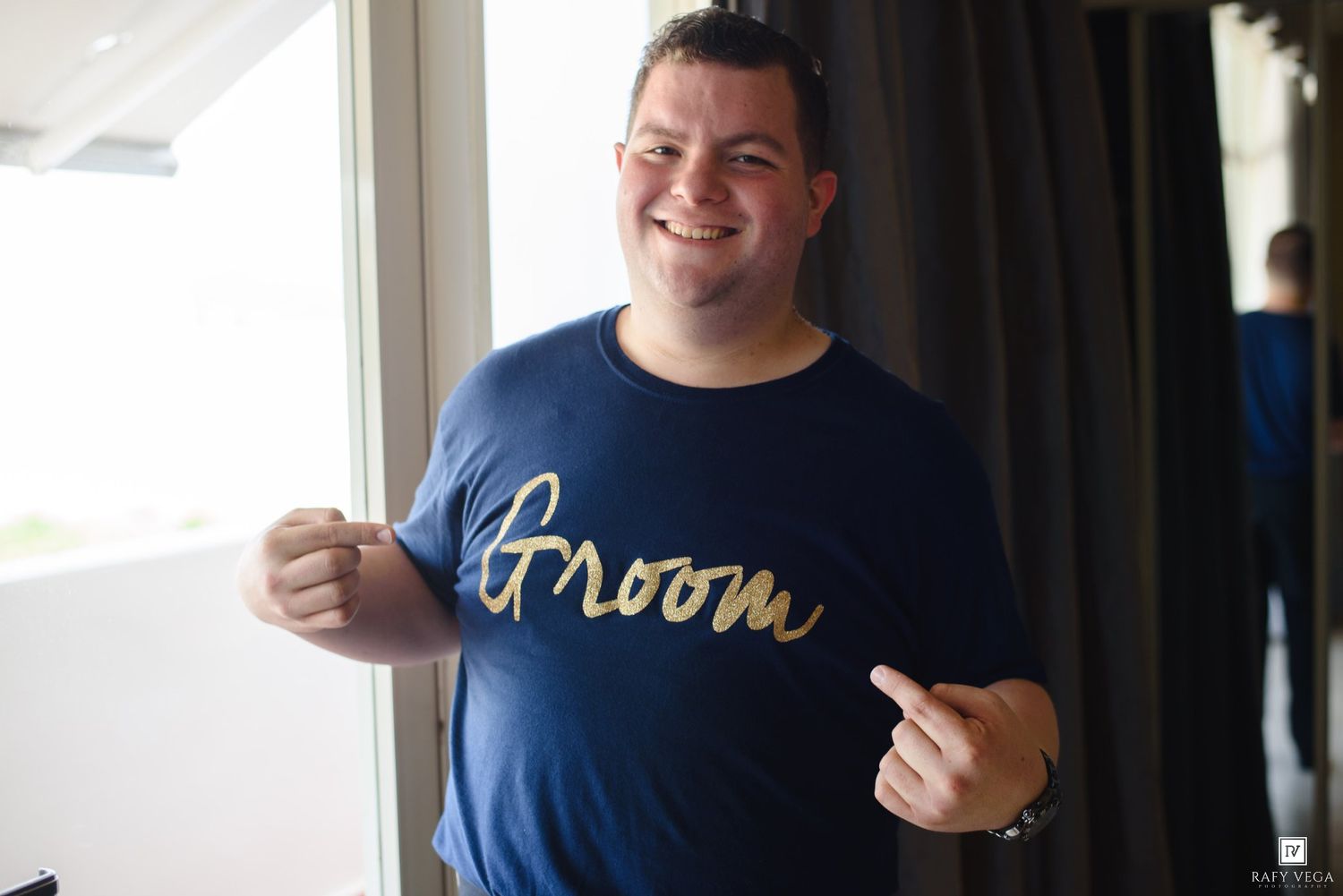 Smiling groom wearing a navy blue Groom t-shirt, pointing and posing happily on his wedding day.