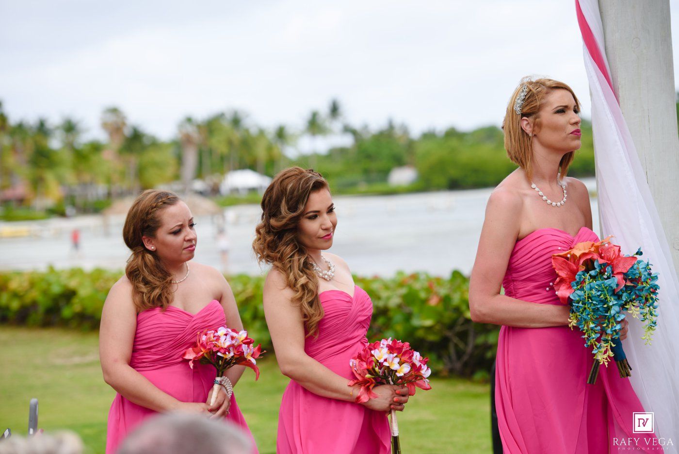 Three bridesmaids in pink strapless dresses holding colorful bouquets stand at an outdoor tropical wedding ceremony.