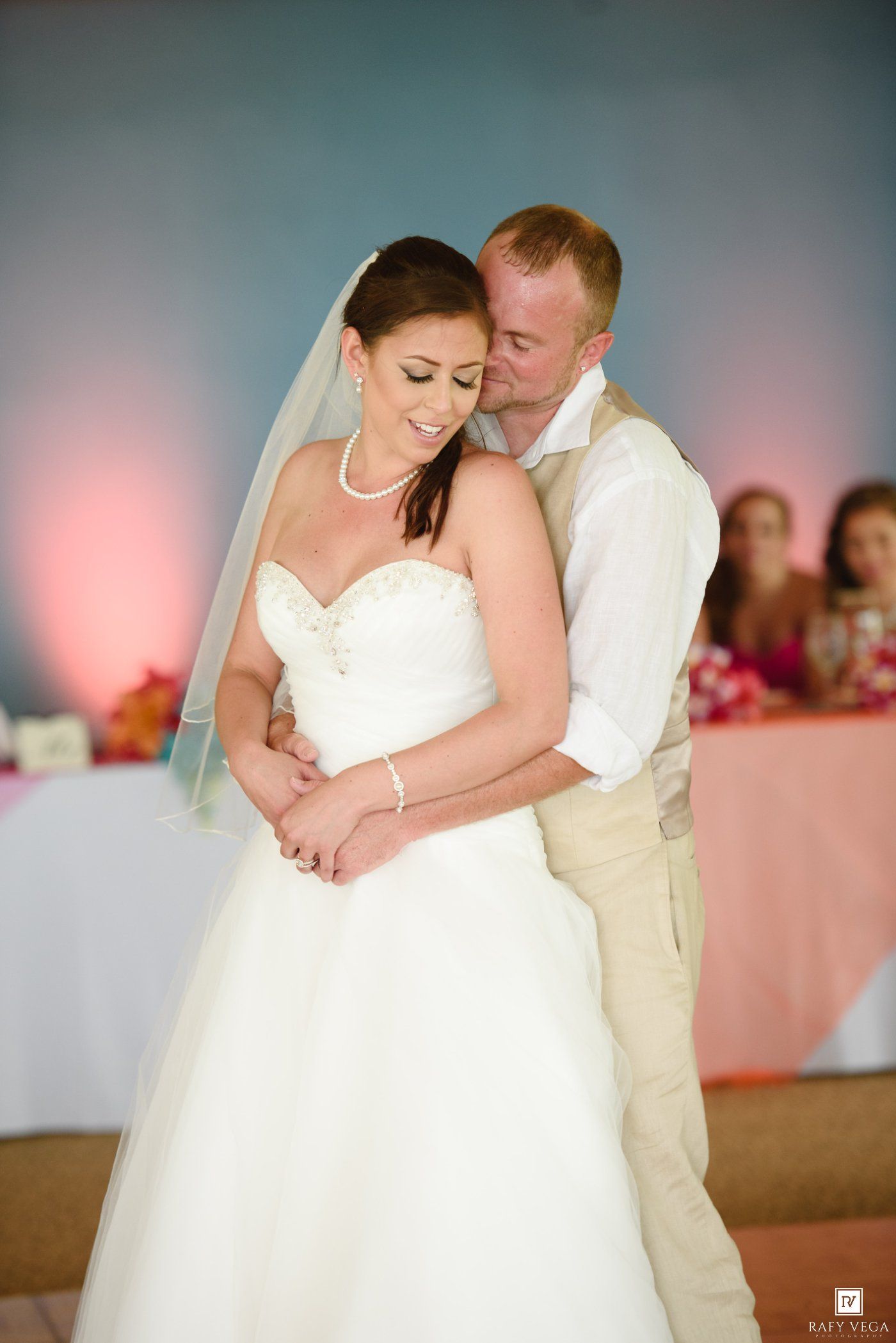 Bride and groom share an intimate first dance, with groom embracing bride in white strapless gown and veil at wedding reception.