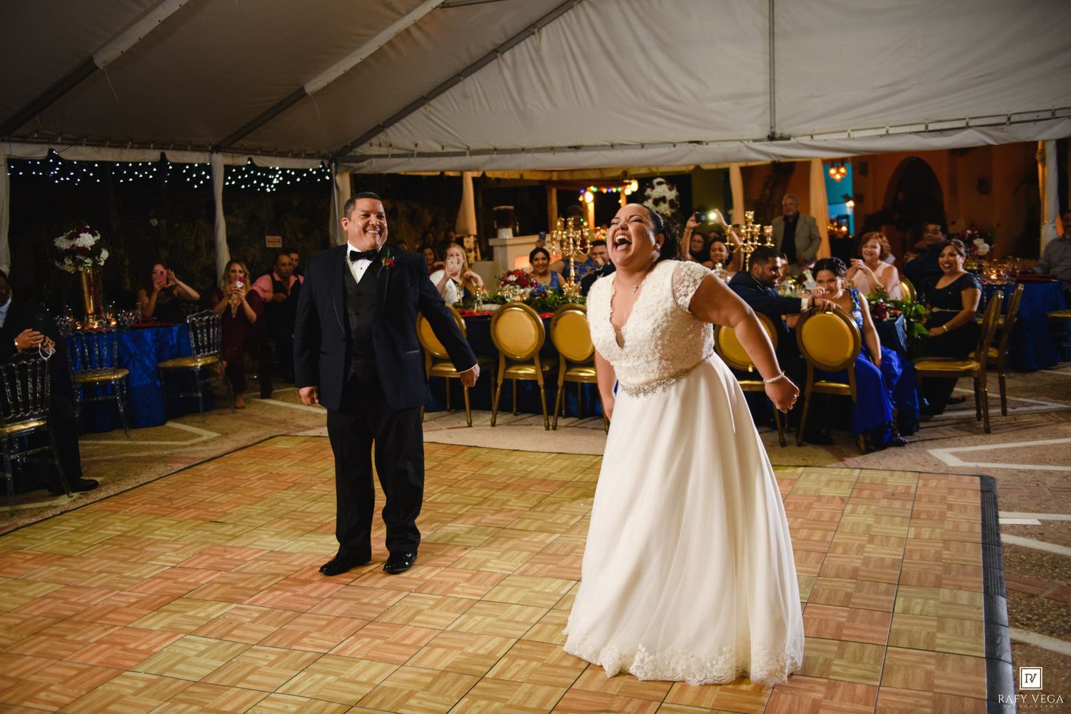 Bride and groom share first dance under elegant tent at wedding reception with guests seated at blue-draped tables.