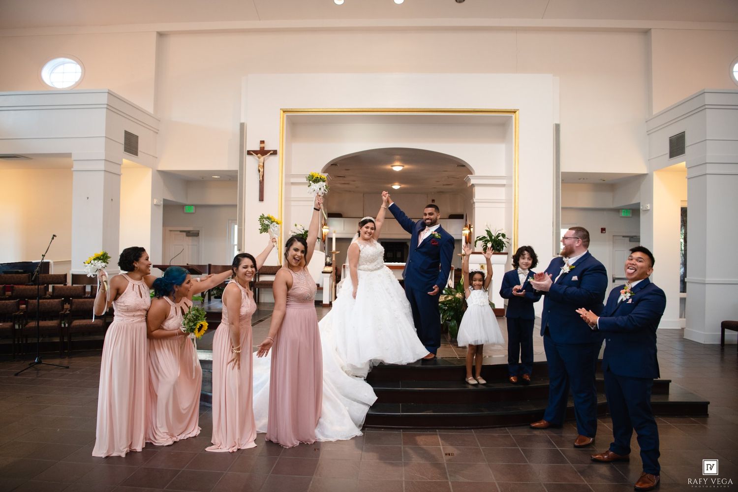 Wedding party celebrating in a church, bridesmaids in blush pink gowns and groomsmen in navy suits cheering for the newlyweds.