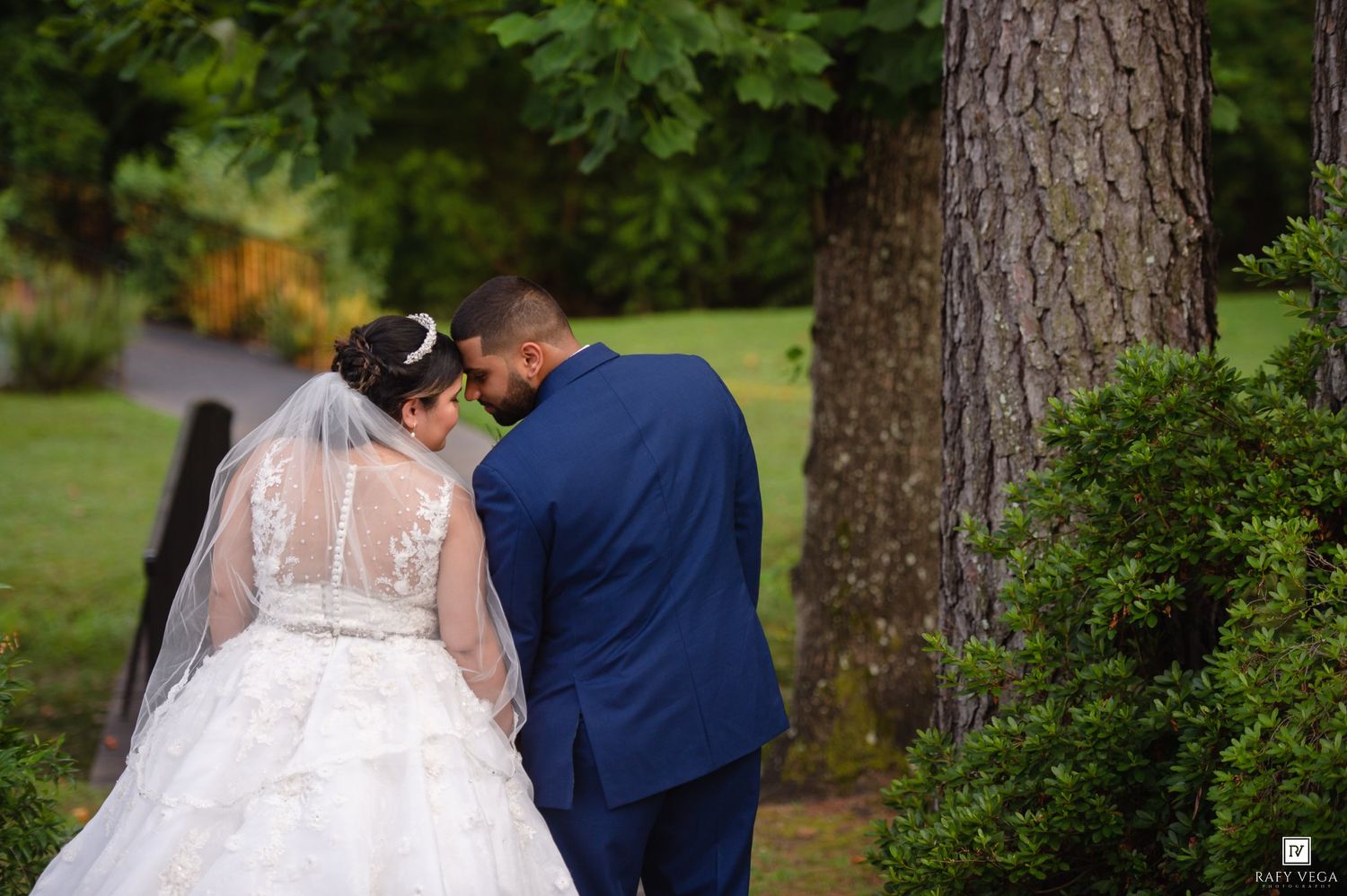 Bride in white gown and veil with groom in navy suit sharing intimate moment near large tree in lush garden.