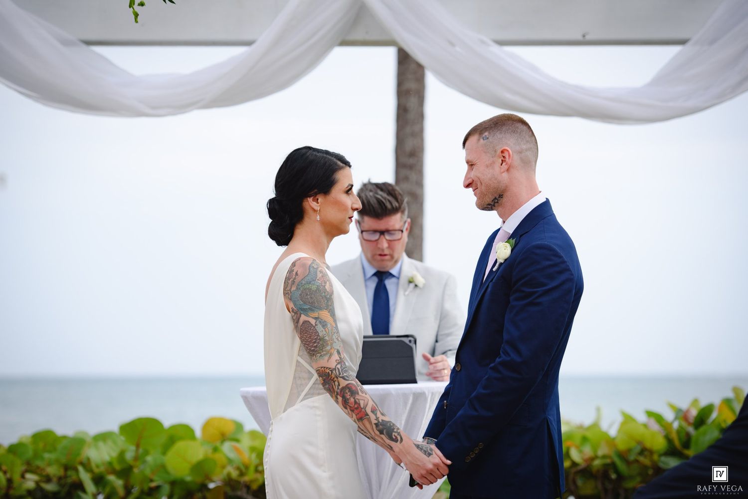 Couple exchanges vows at outdoor beach wedding ceremony with officiant and ocean backdrop.
