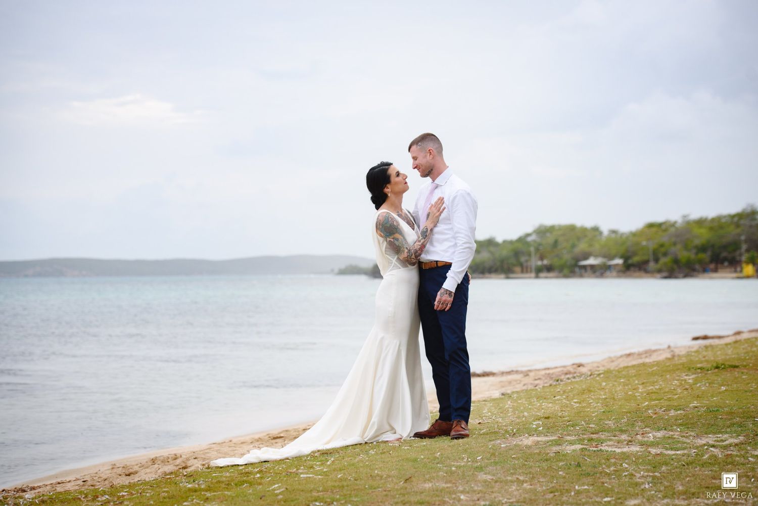 Bride and groom share a romantic moment on a beach shoreline, bride in elegant white gown, groom in white shirt and navy pants.
