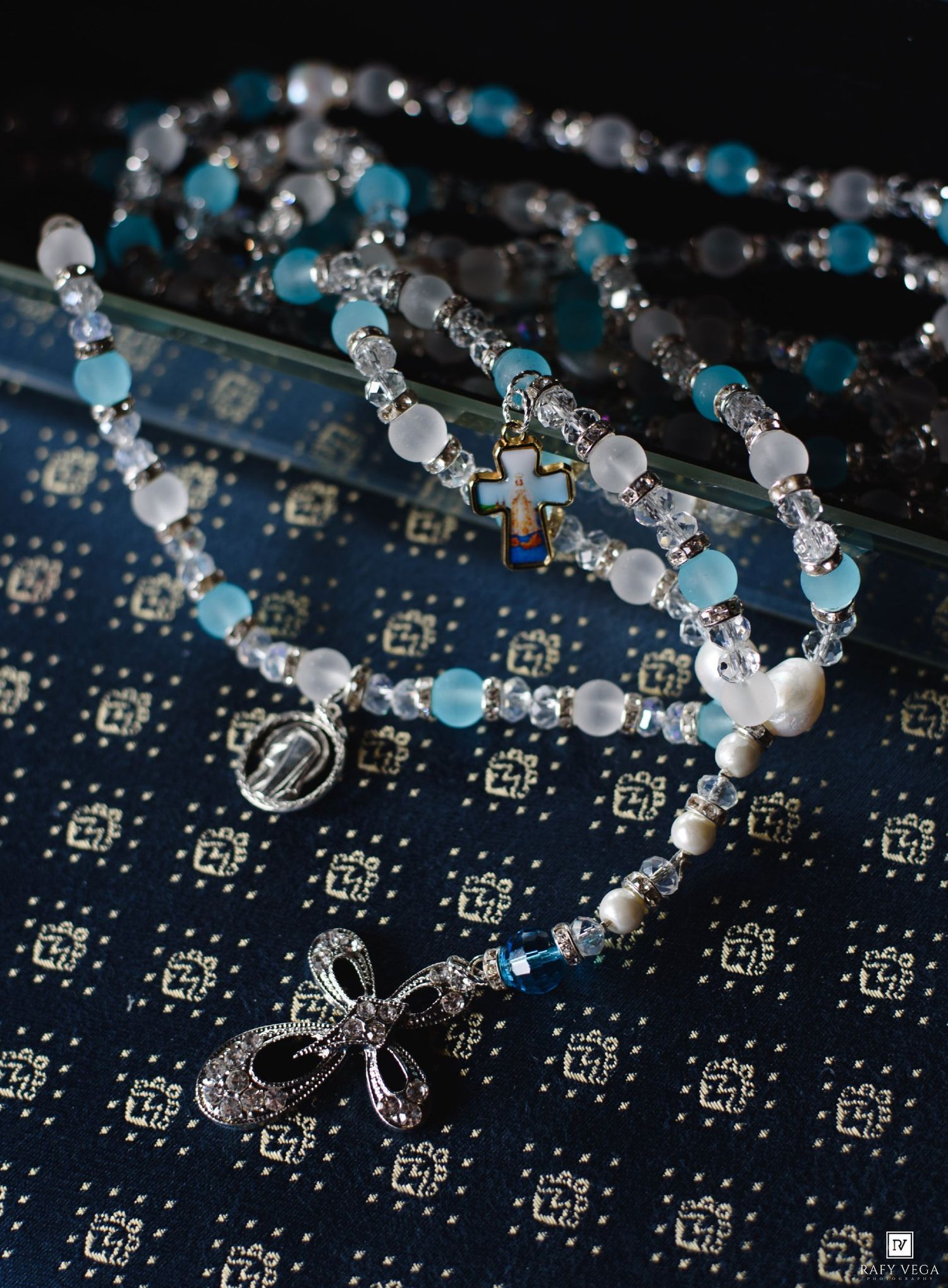 Blue and white beaded rosary necklaces with cross and bow pendants displayed on decorative fabric.