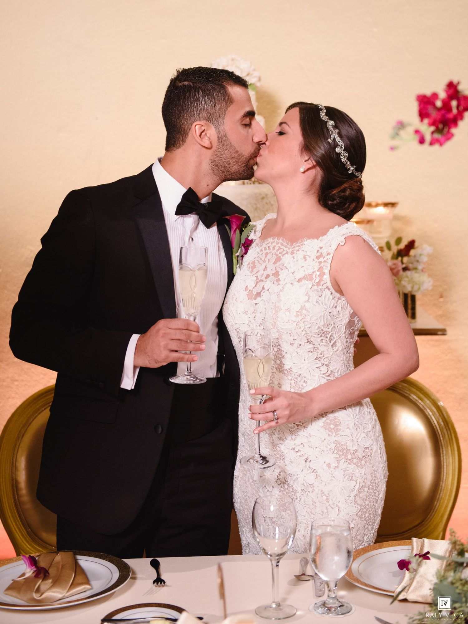 Bride and groom share a kiss while holding champagne glasses at their elegant wedding reception dinner table.