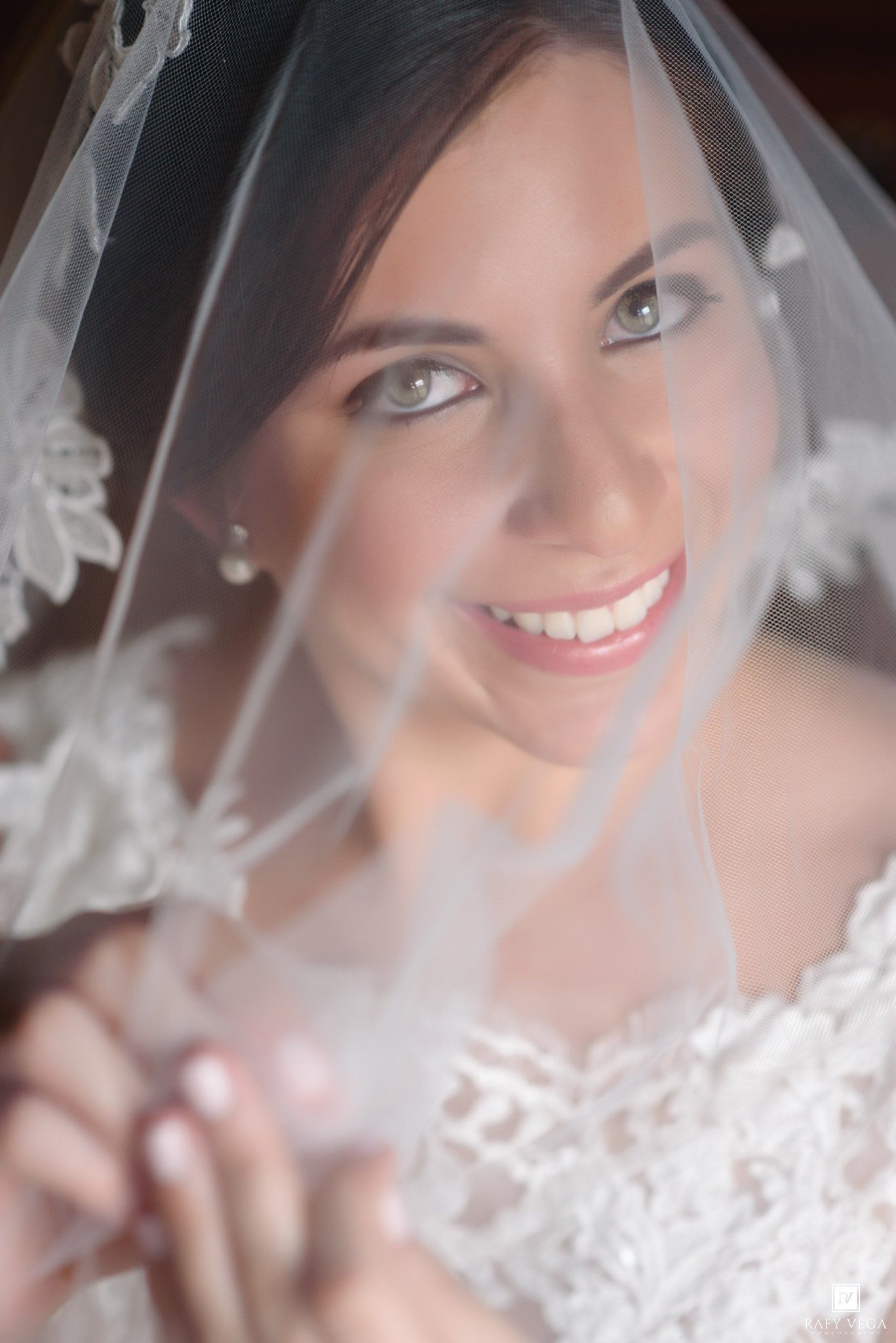 Smiling bride wearing lace veil and earrings holds veil forward on her wedding day.