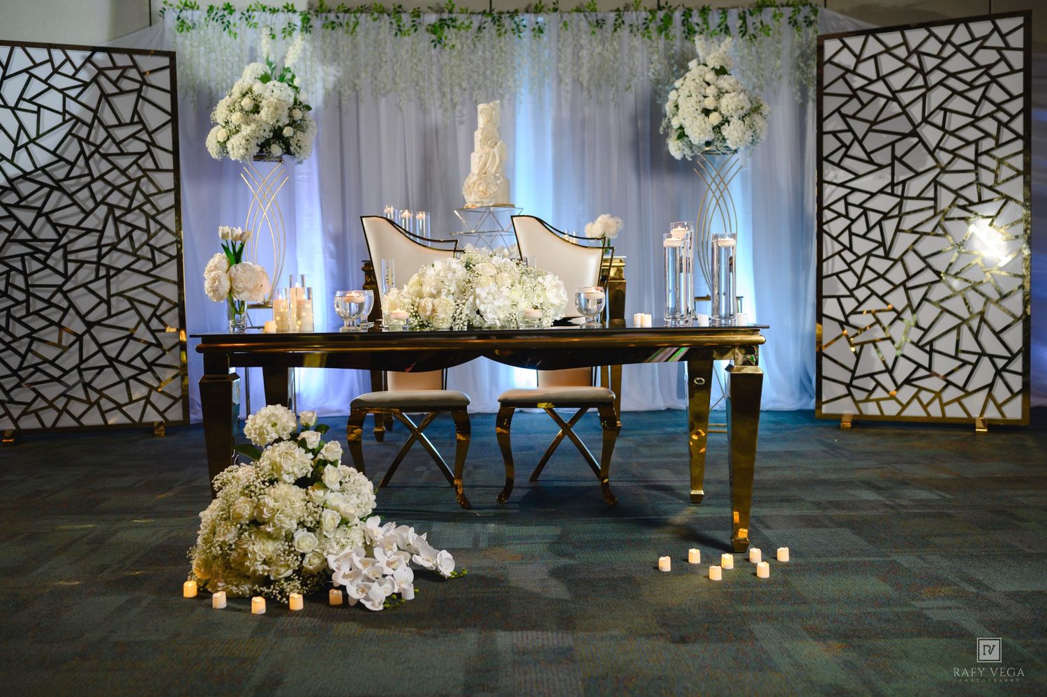 Elegant wedding sweetheart table with gold accents, white floral arrangements, candles, and decorative panels in a banquet hall.