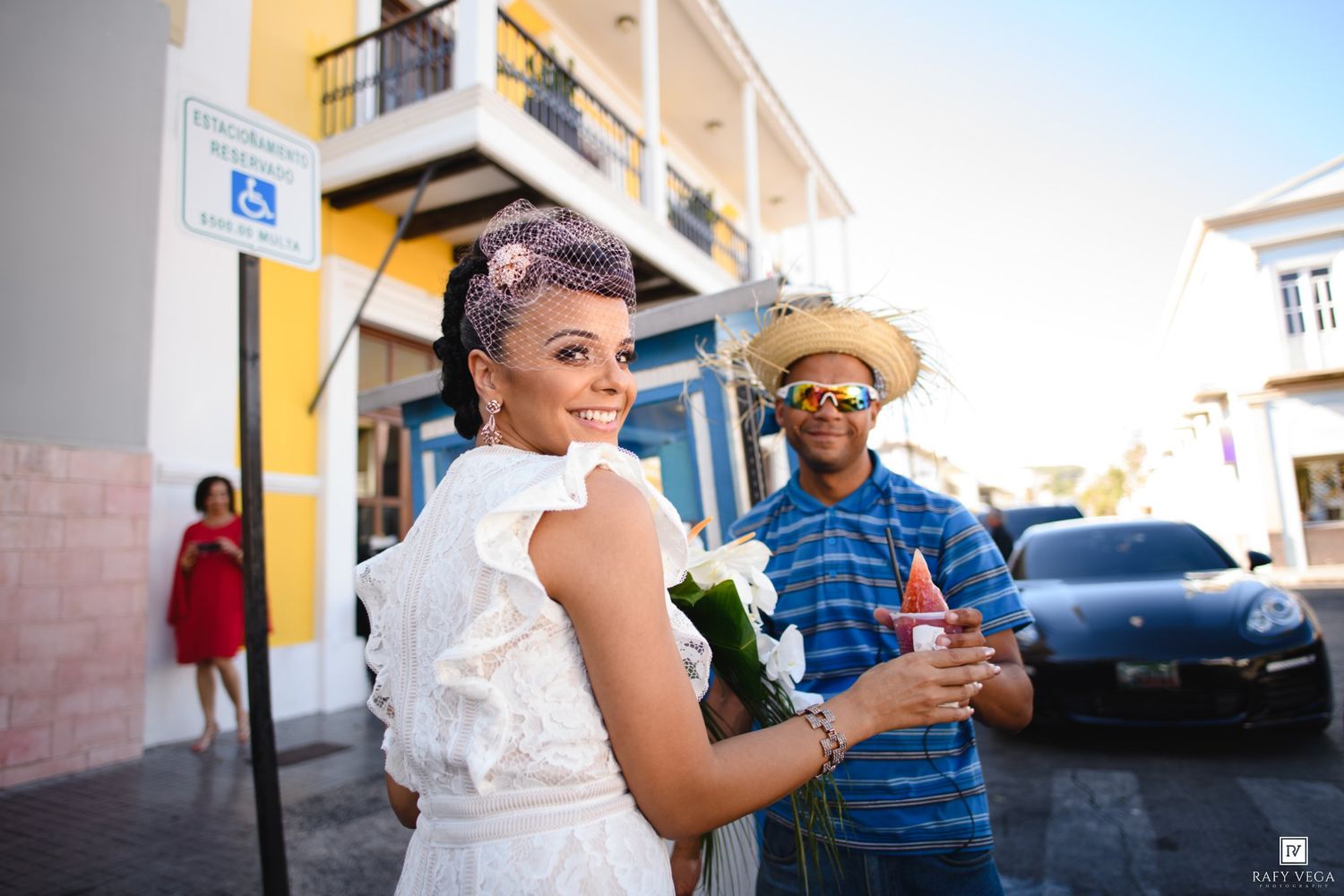 A woman in a white dress holds flowers while a man in a straw hat enjoys a treat on a colorful street.