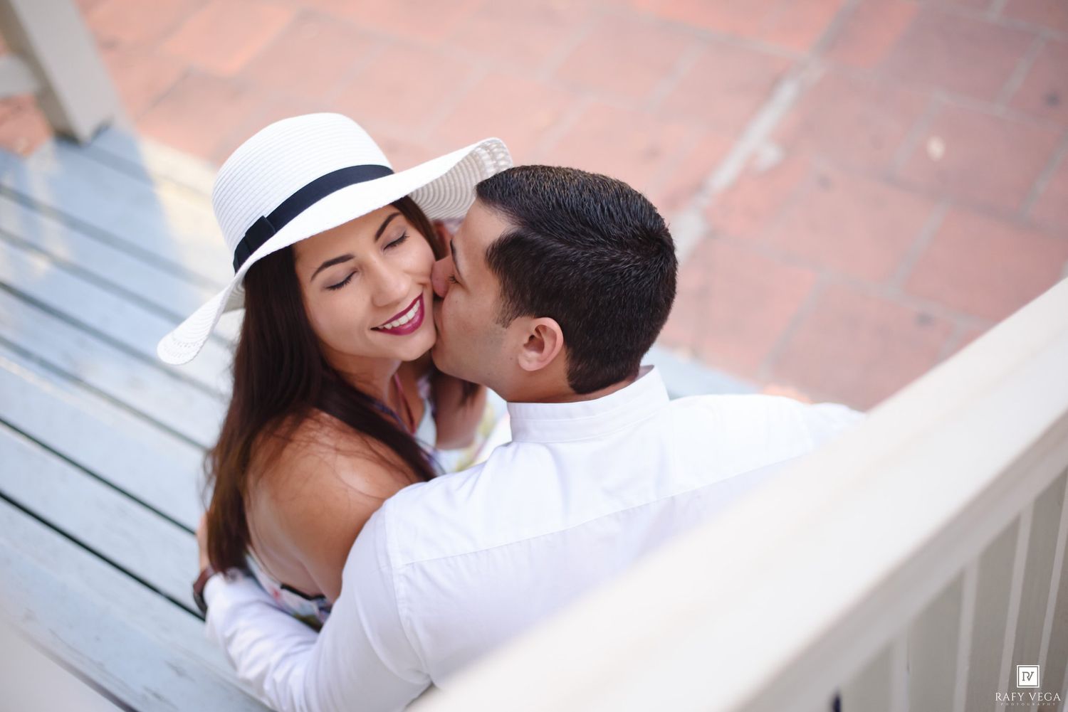 A couple shares a romantic kiss on sun-drenched stairs, woman in white hat smiling joyfully.