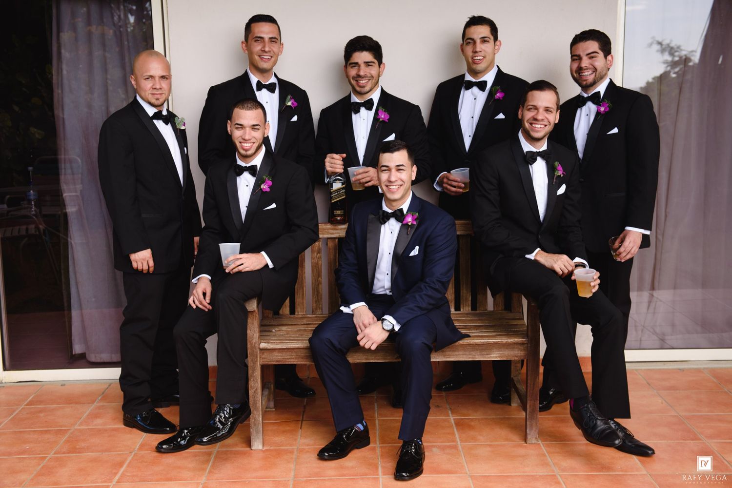 Groom and groomsmen in black tuxedos with pink boutonnieres posing together at a wedding celebration.