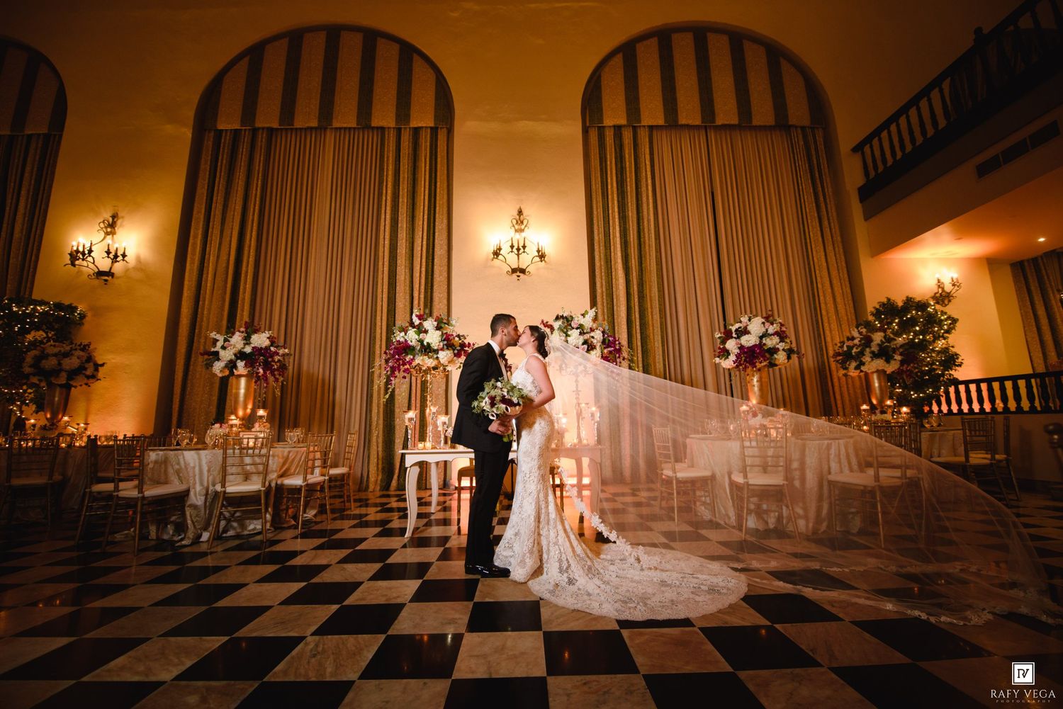Bride and groom share a romantic kiss in an elegant ballroom with checkered floors and warm golden lighting.