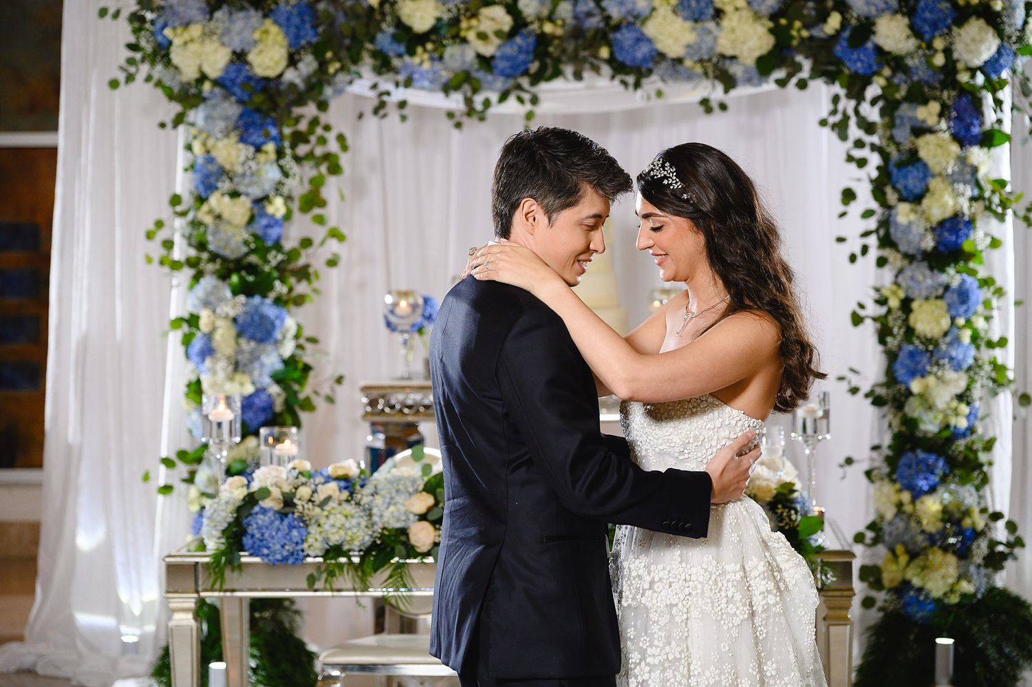 A bride and groom share a romantic first dance under a floral arch adorned with blue and white flowers at their wedding.