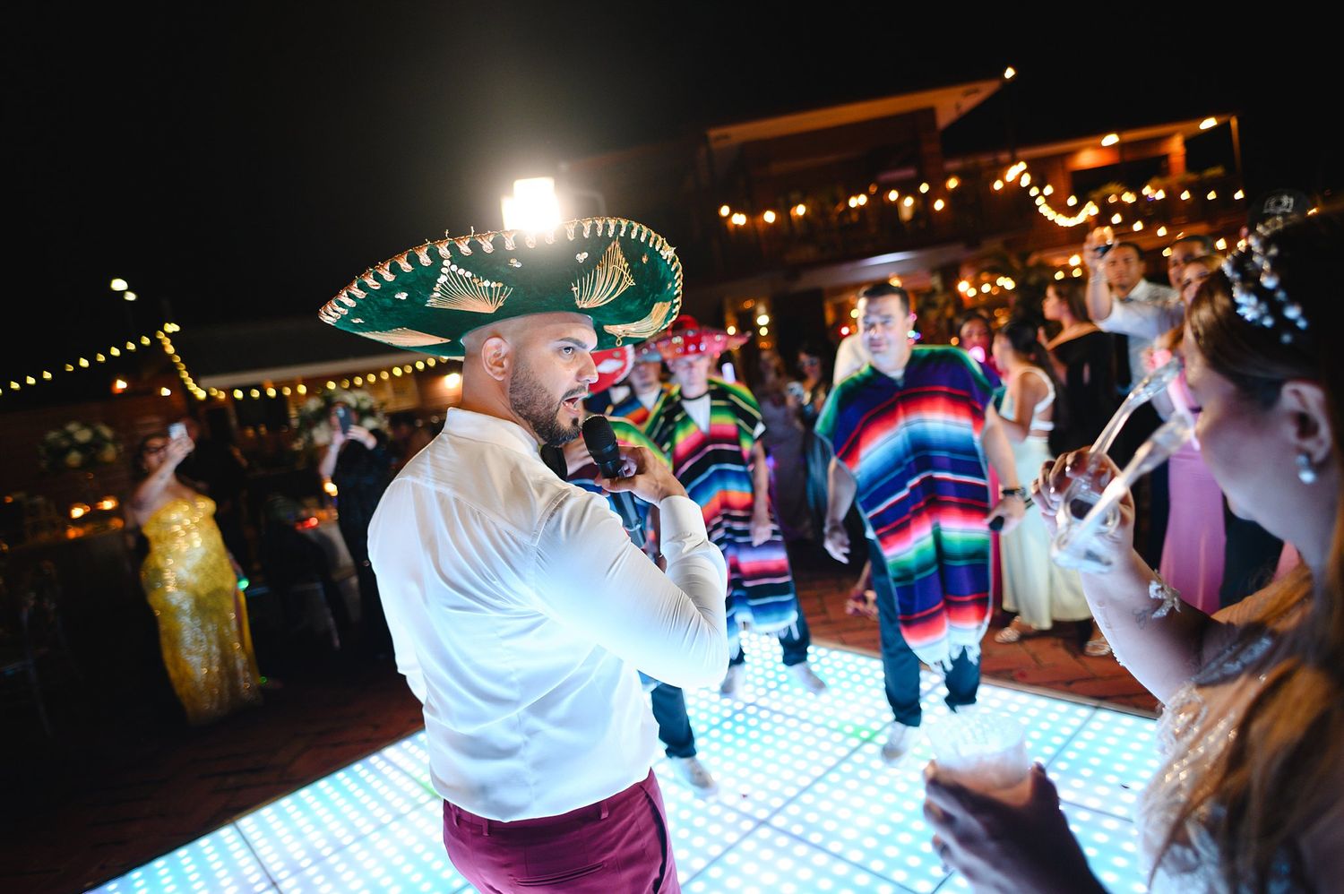 Festive Mexican fiesta wedding reception with guests dancing, wearing sombreros and colorful ponchos on illuminated dance floor.