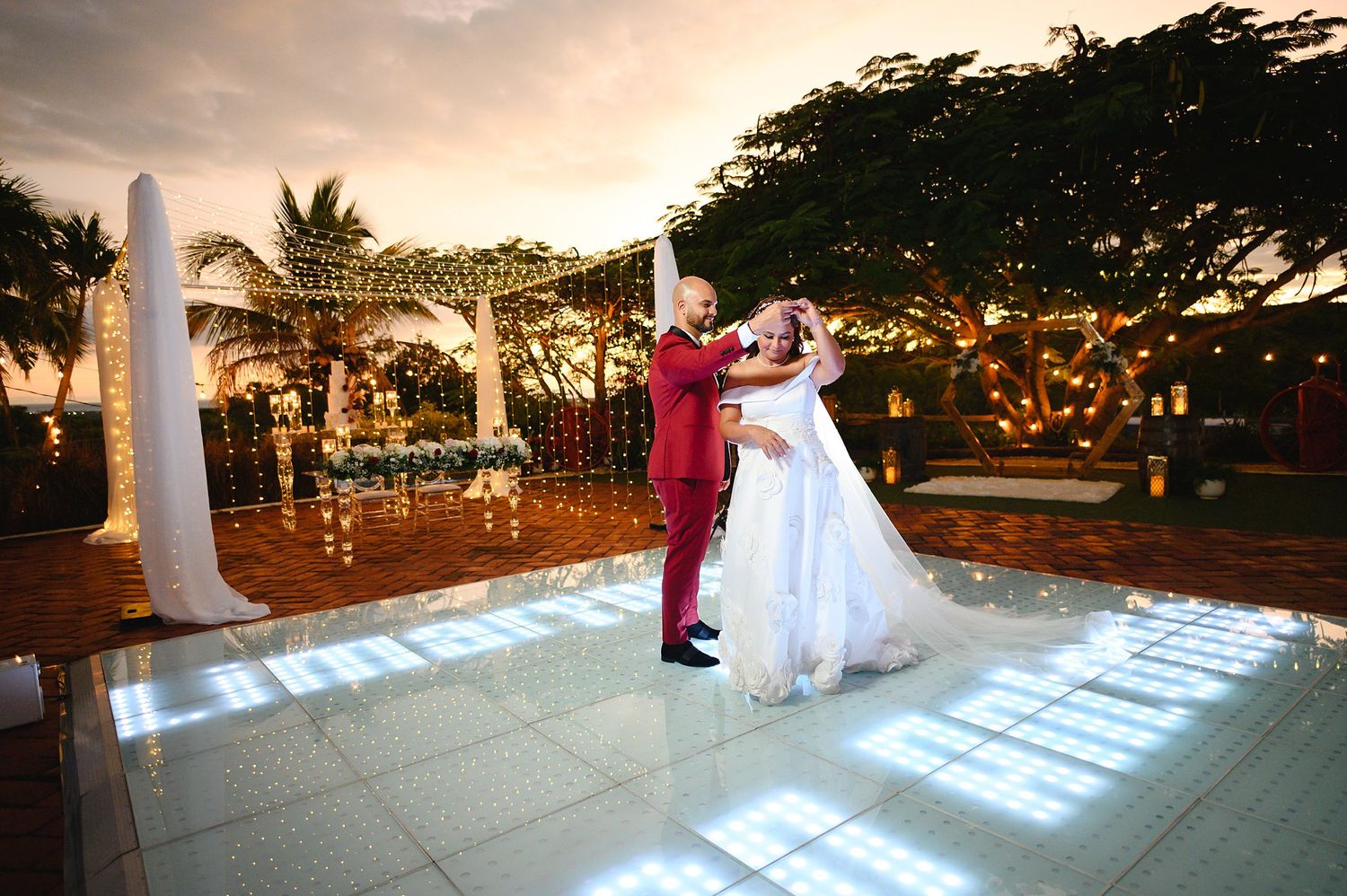 Bride and groom share first dance on illuminated outdoor floor at sunset wedding reception.