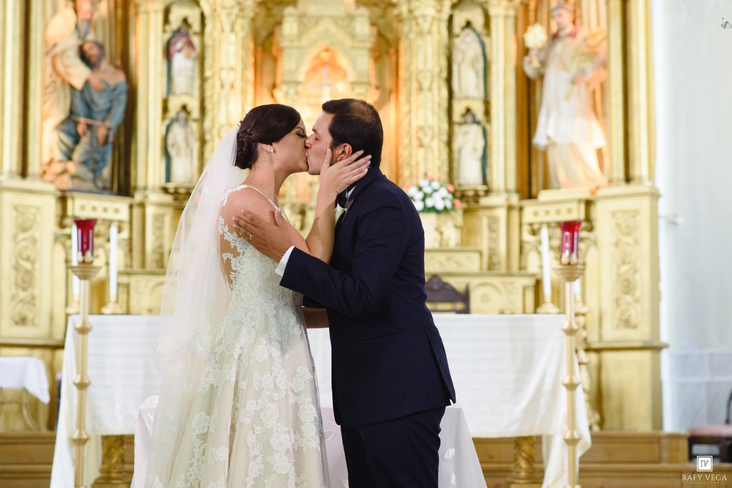 Bride and groom share first kiss at ornate golden church altar during traditional wedding ceremony.