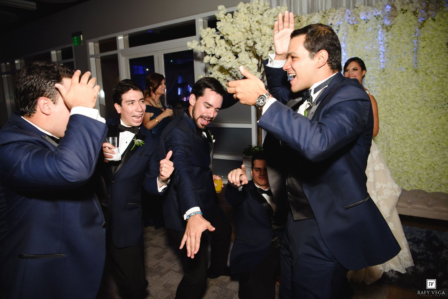 Groomsmen in navy suits celebrate joyfully at a wedding reception, dancing and high-fiving near floral decorations.
