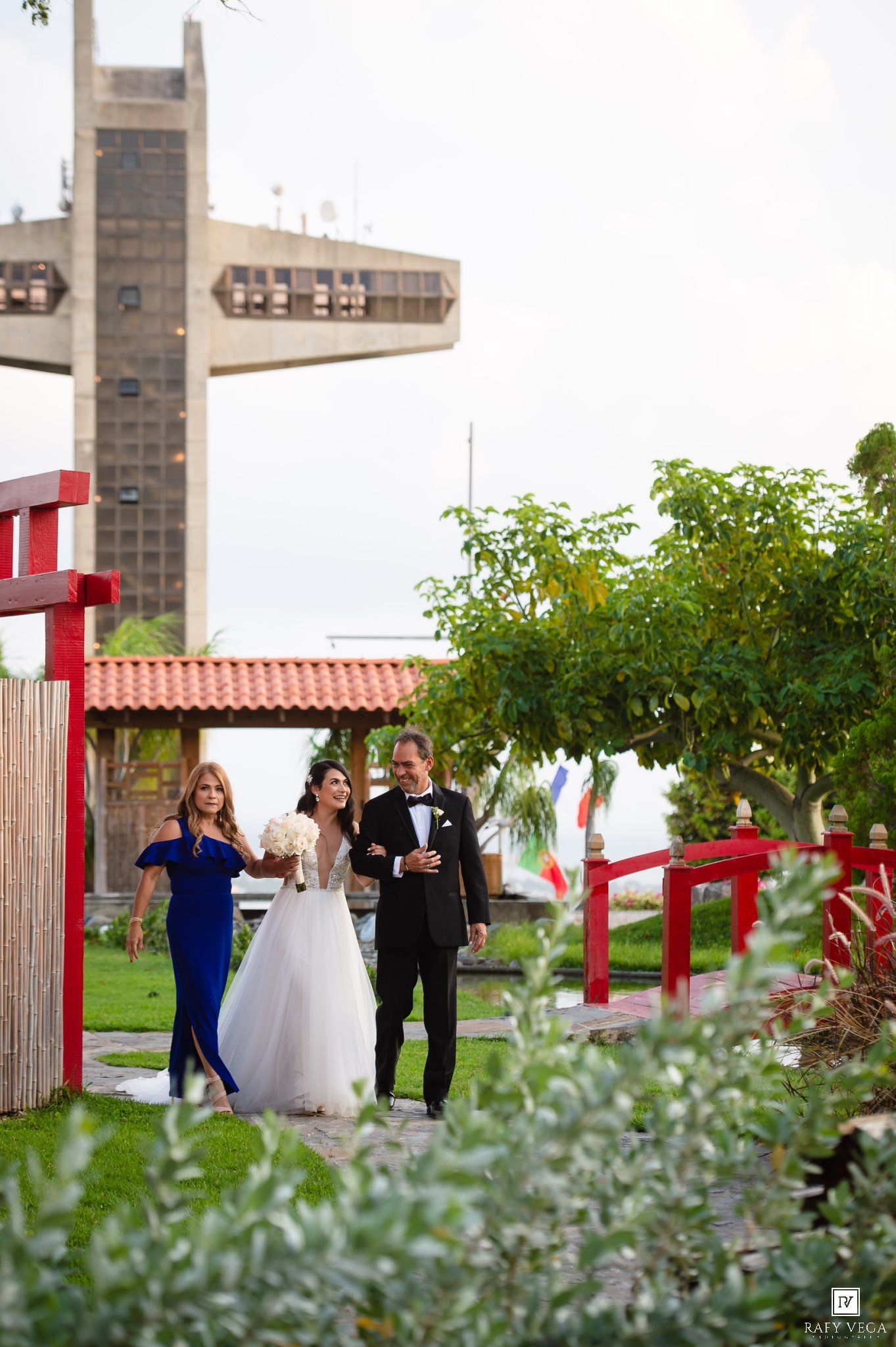 Bride in white gown walks with groom in tuxedo and woman in blue dress through a garden wedding venue.