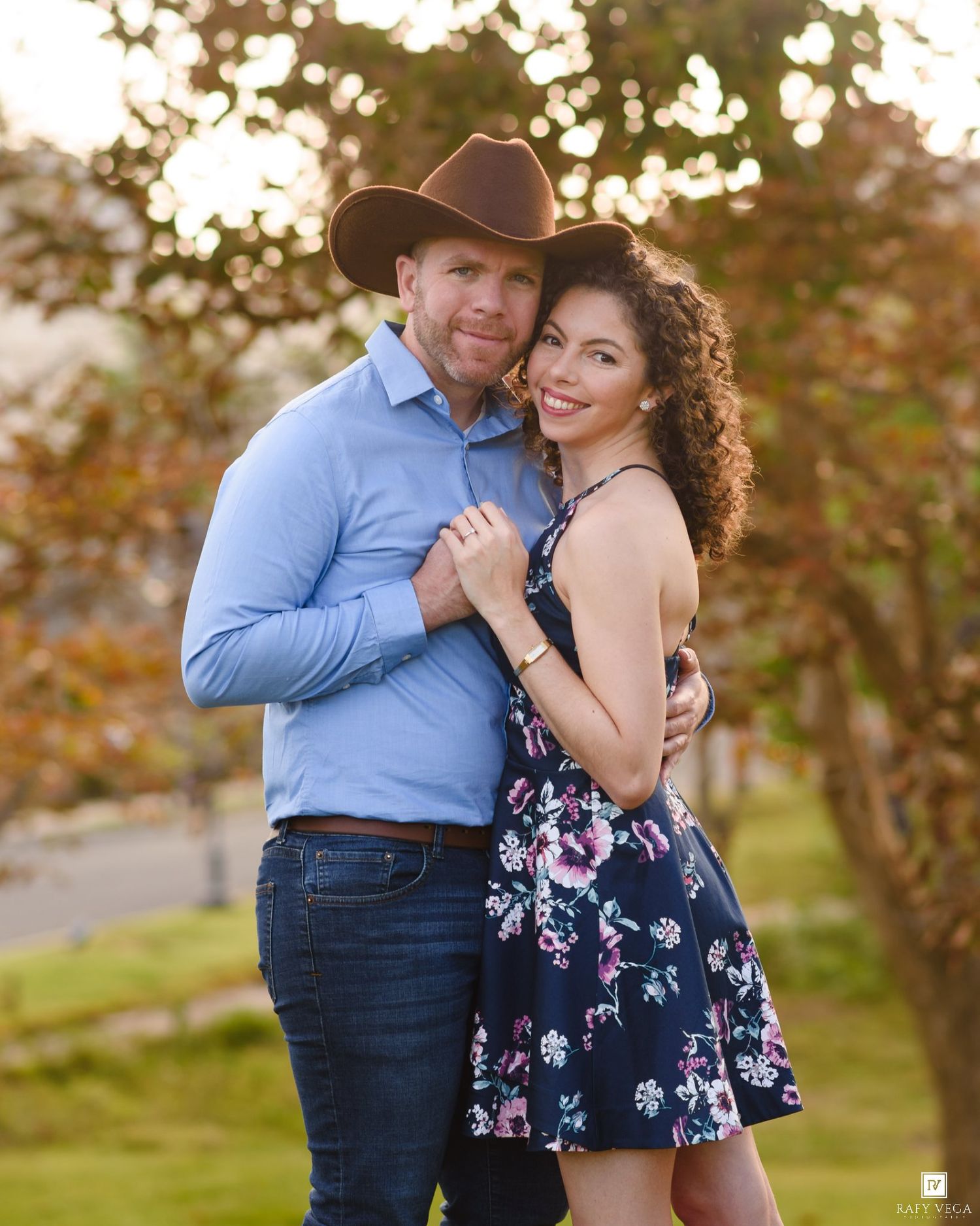 Couple posing outdoors, man in cowboy hat and blue shirt, woman in floral dress, embracing near autumn trees.