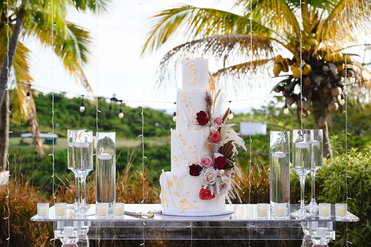 Elegant white multi-tier wedding cake with red roses and gold accents displayed outdoors with tropical palm trees.