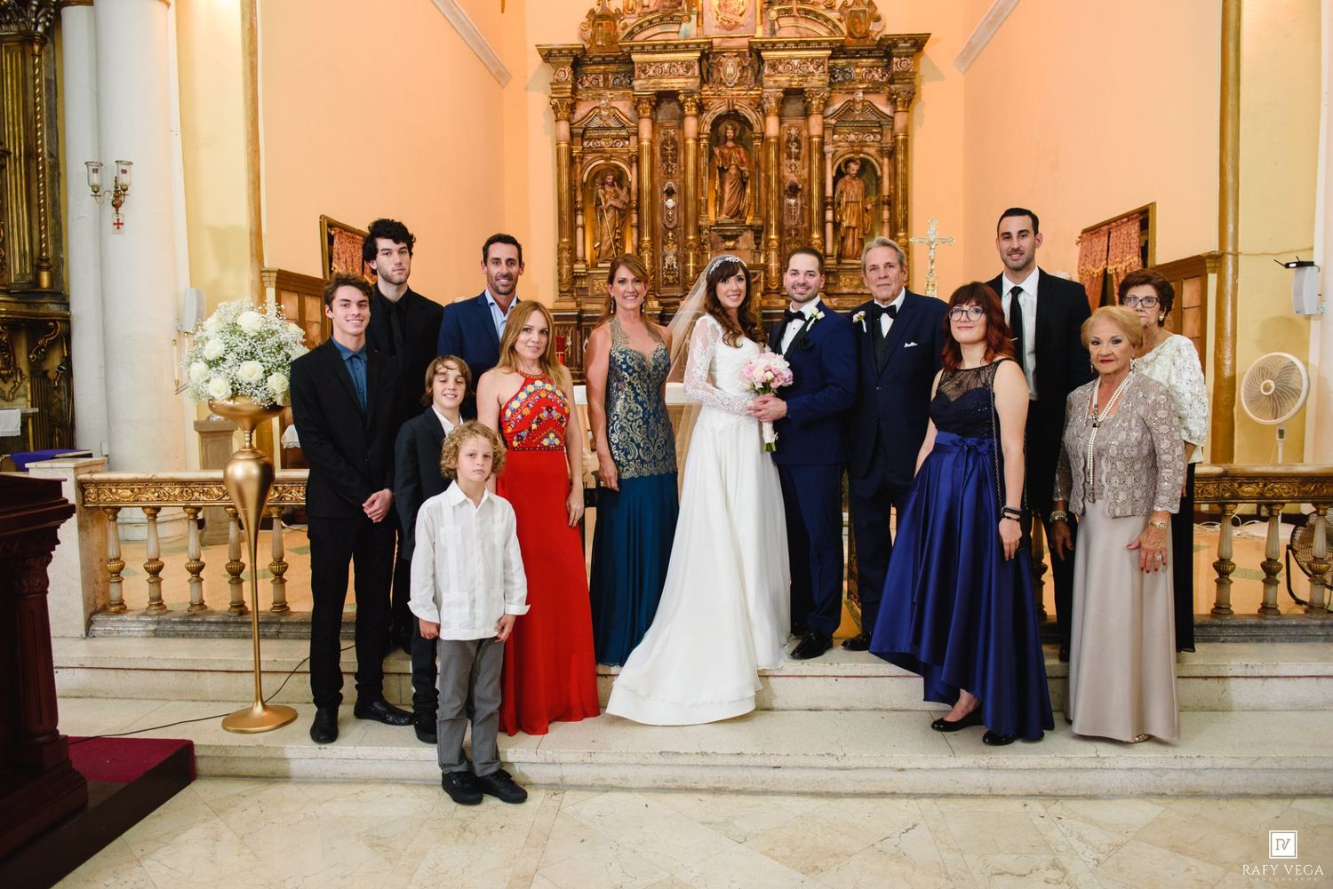 Wedding party posing together inside an ornate church with golden altar, bride in white gown surrounded by family.
