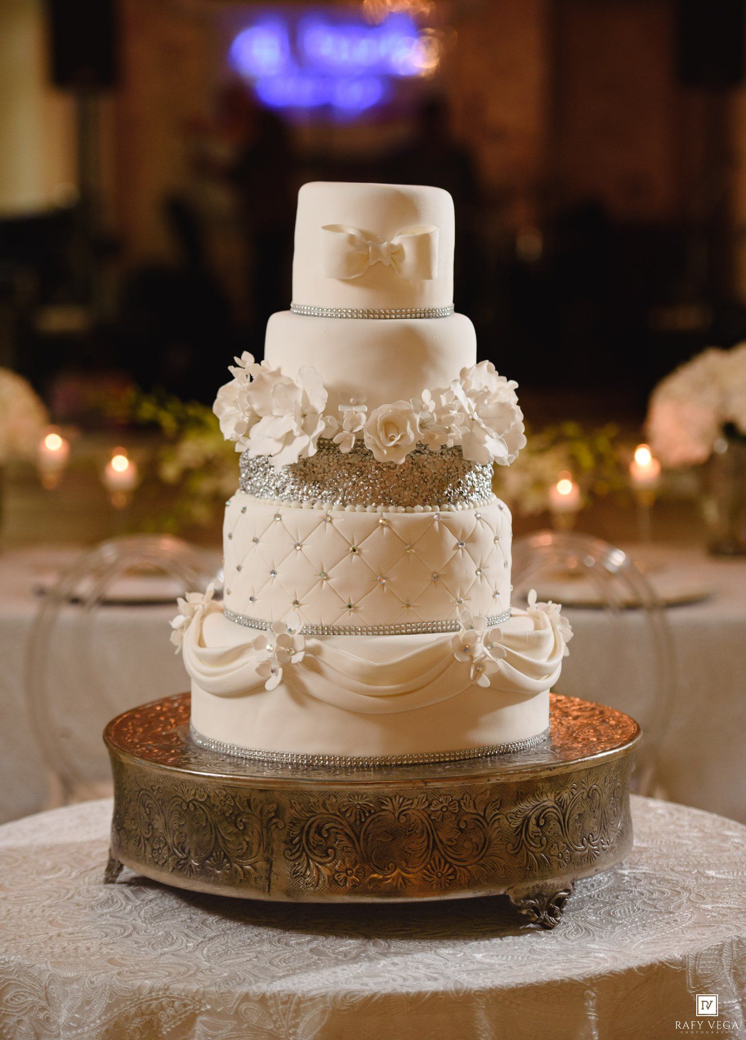Ornate four-tier white wedding cake with quilted details, rhinestones, and floral accents on a gold stand.