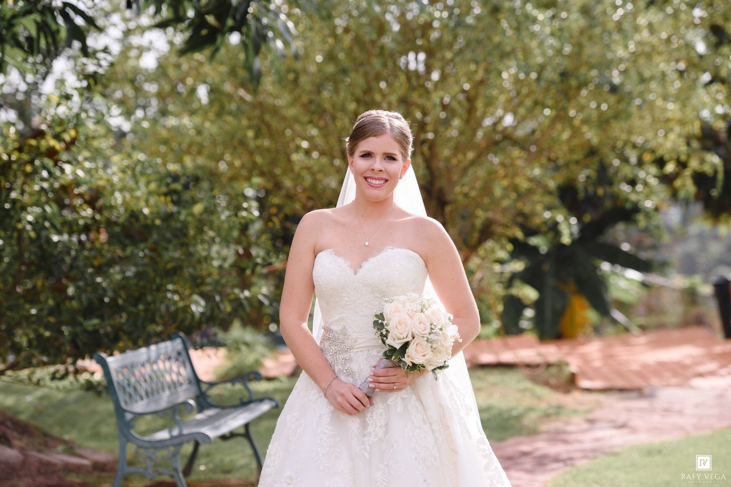 Bride in strapless lace gown holds white bouquet outdoors on wedding day.