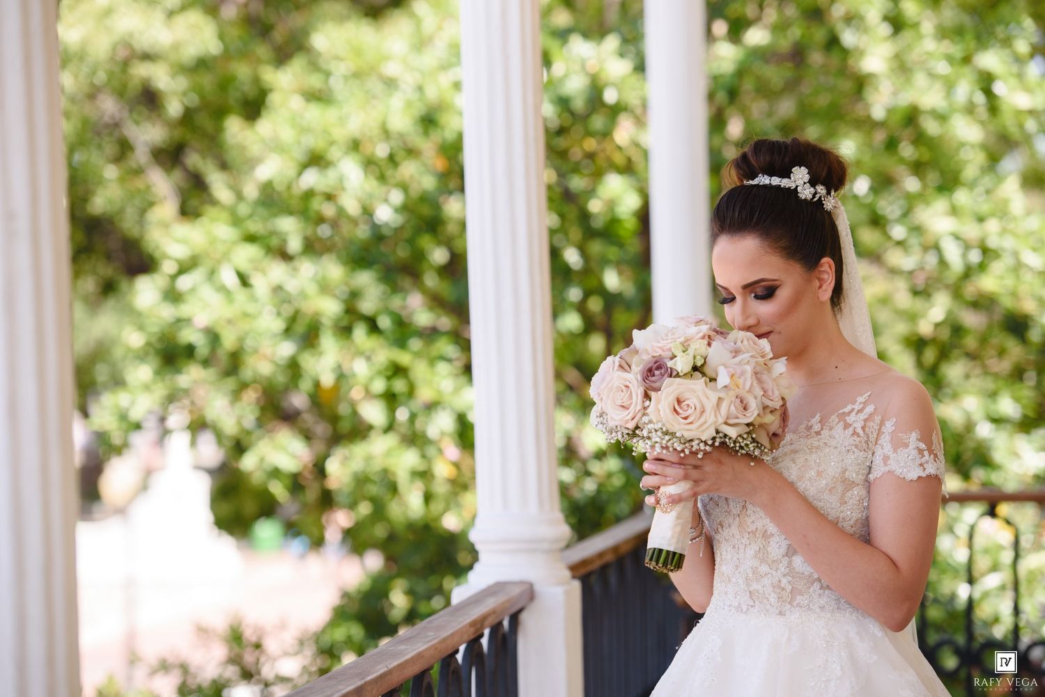 Bride in lace wedding gown smelling bouquet of blush roses on elegant white columned balcony.