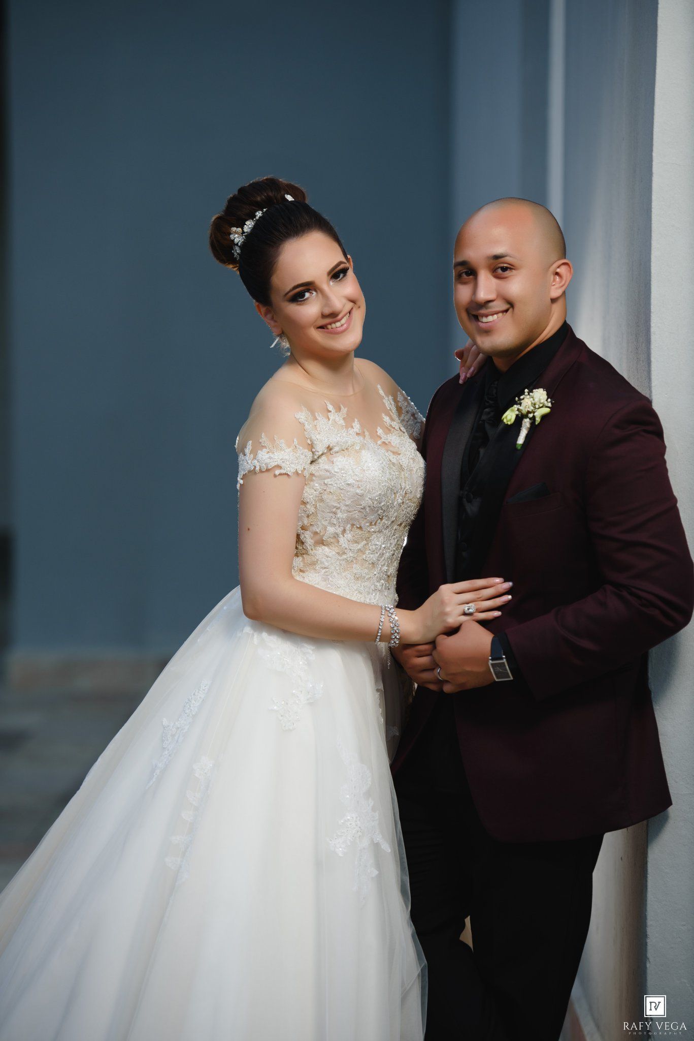 Bride in lace ball gown and groom in burgundy suit pose together smiling at their elegant wedding ceremony.