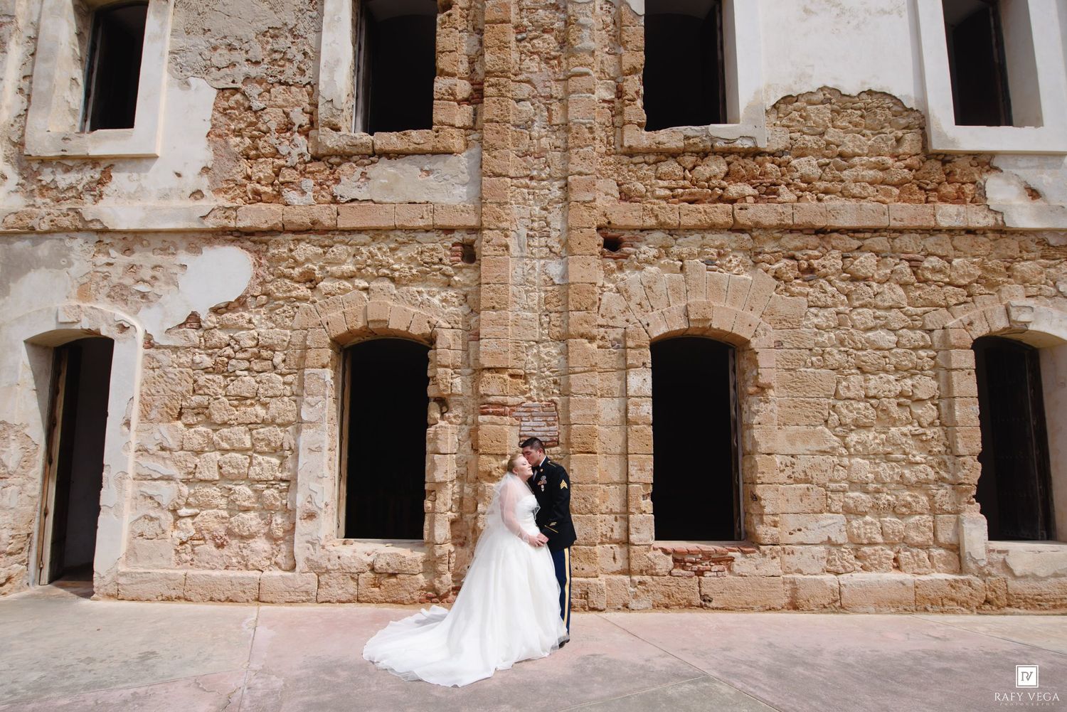 Bride and groom share a romantic kiss in front of a rustic stone building on their wedding day.