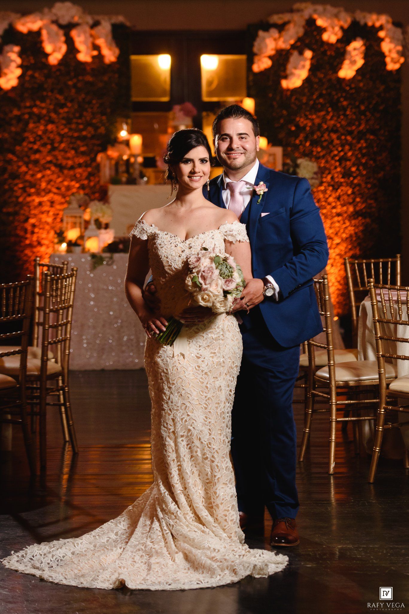 Smiling bride holding floral bouquet poses with groom in navy suit at warmly lit wedding reception hall.