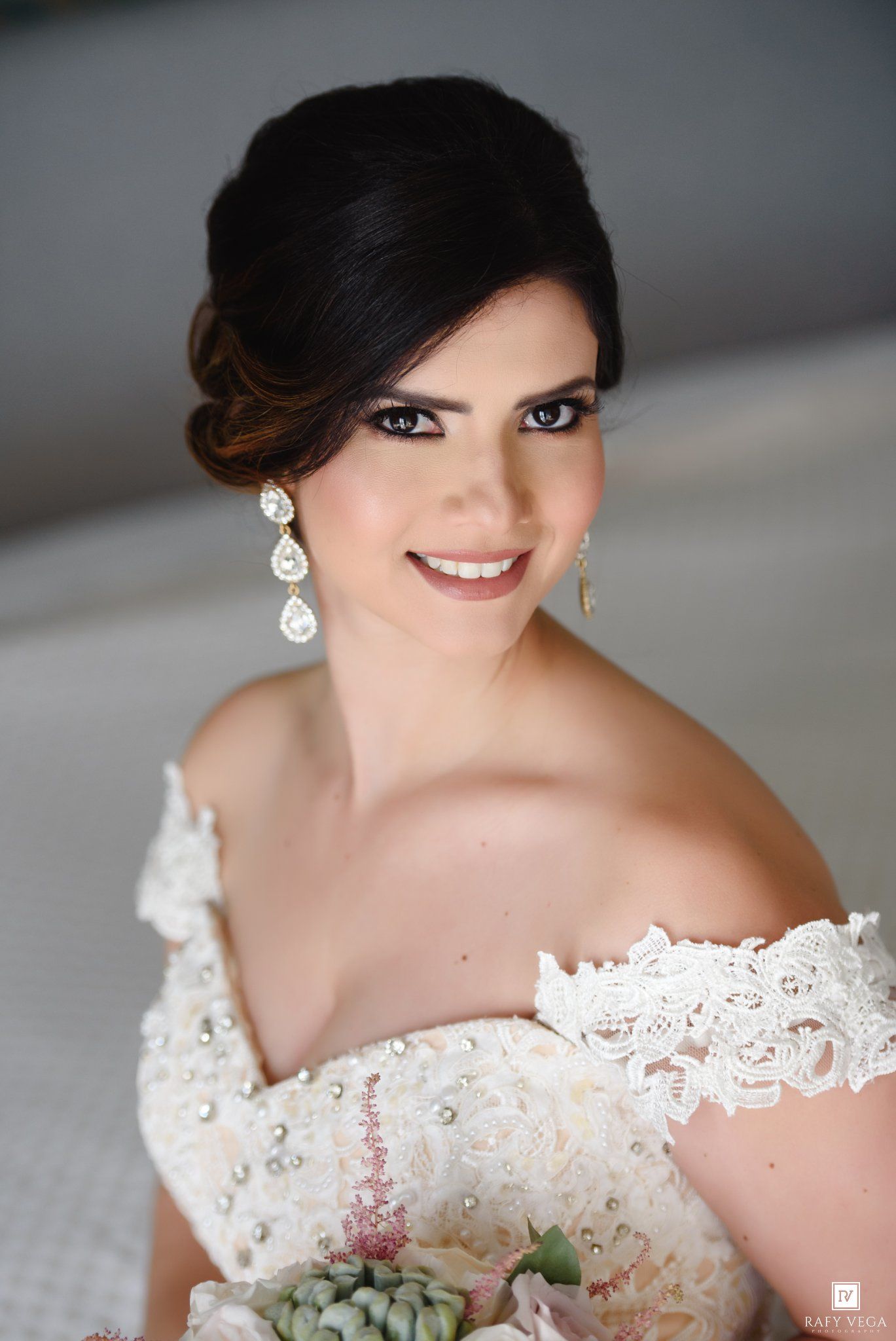 Elegant bride in off-shoulder lace gown holding floral bouquet, wearing teardrop earrings with updo hairstyle.