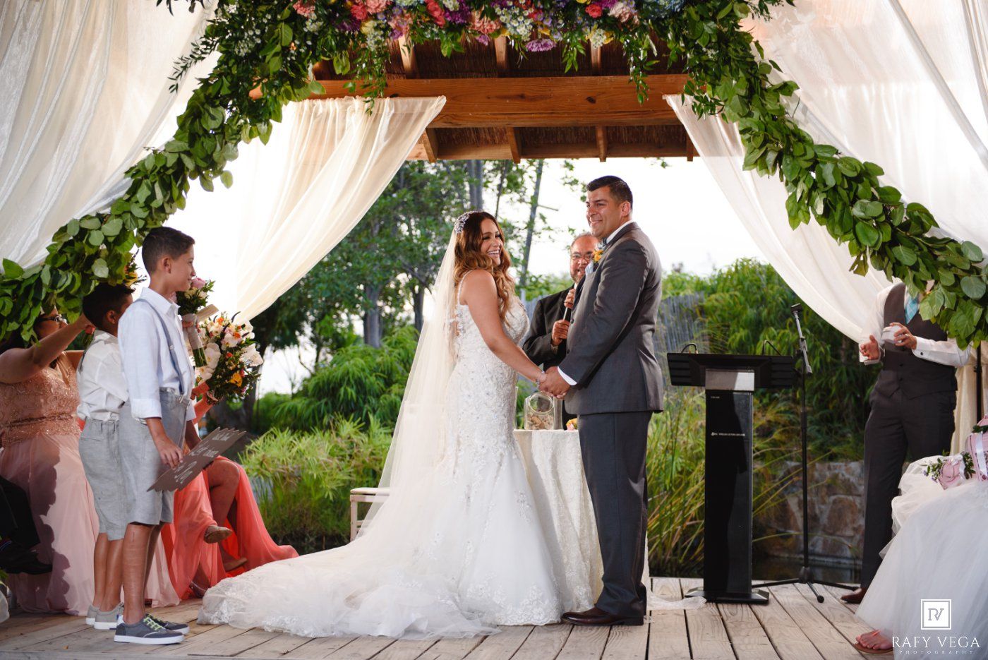 Bride and groom exchange vows at an outdoor wedding ceremony under a floral arch with white draping.