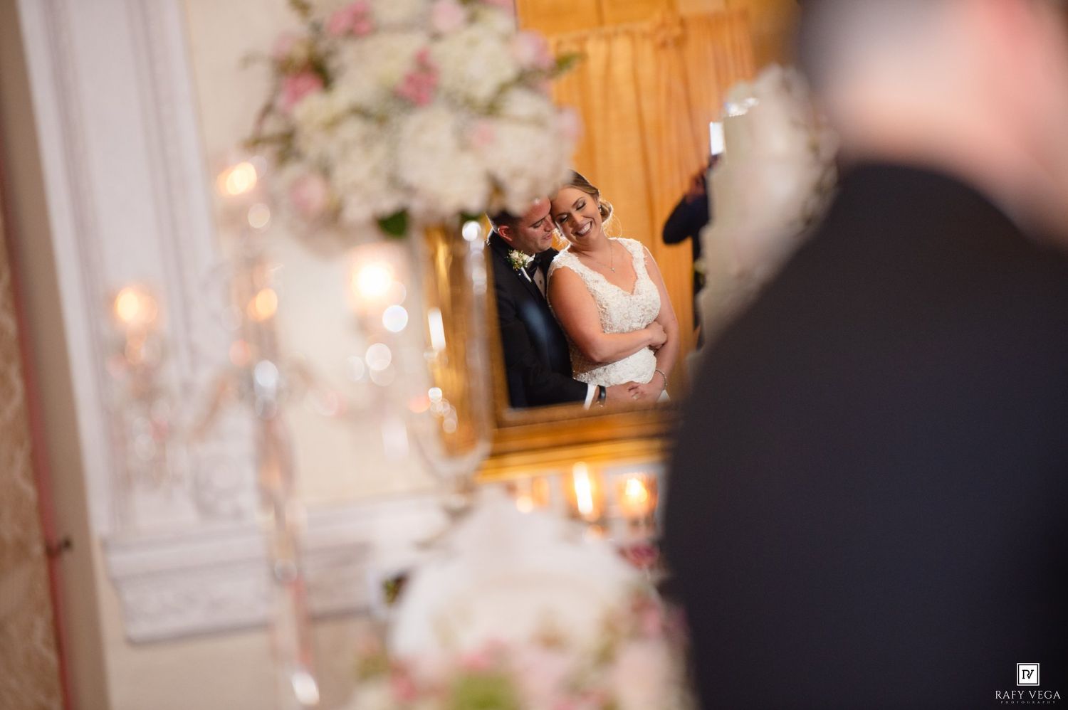 Bride and groom share a romantic moment reflected in an ornate gold mirror surrounded by elegant floral arrangements.