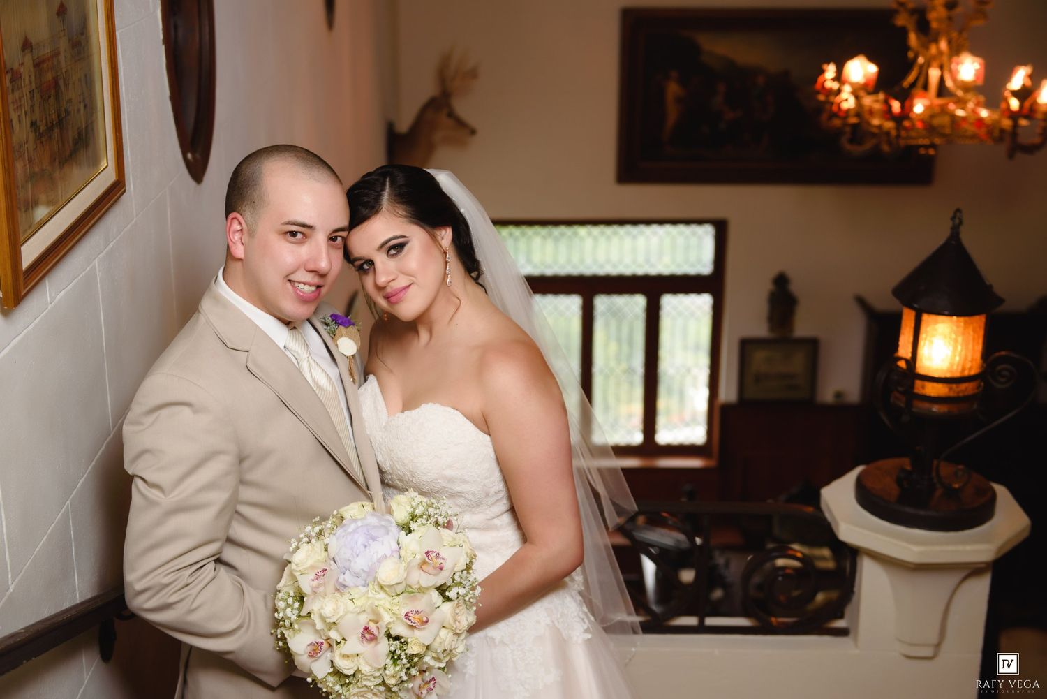 A bride and groom pose together indoors, she holds a white bouquet, wearing a strapless gown, he wears a beige suit.