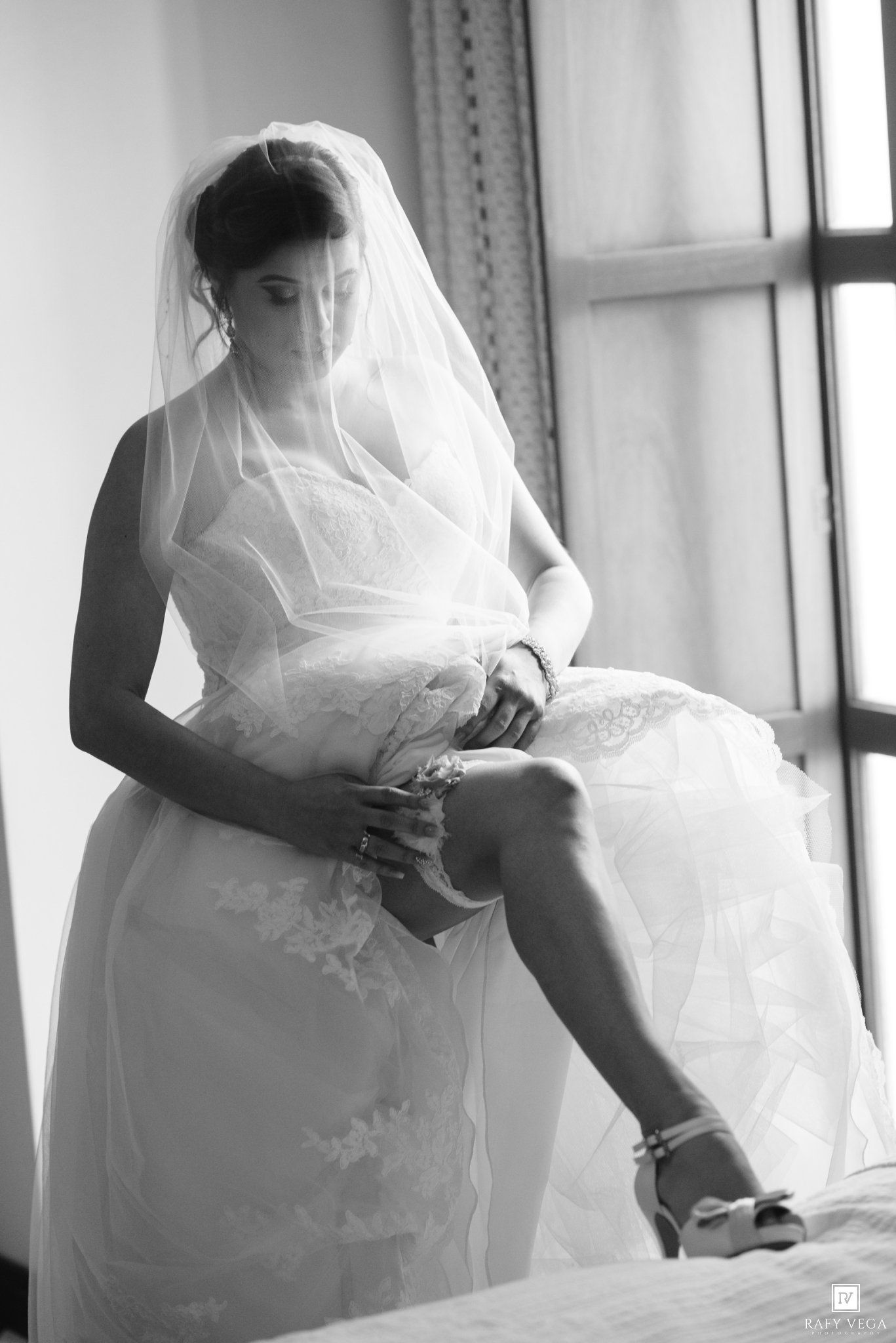 Black and white photo of a bride in a lace gown and veil fastening her heels while seated near a window.