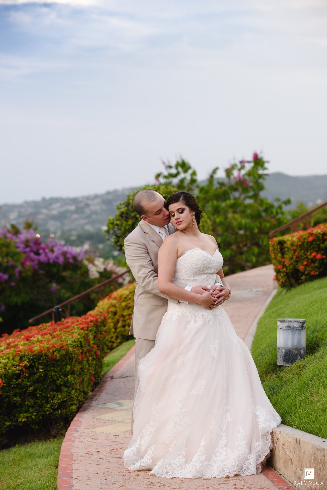 Bride in lace gown and groom in tan suit share a romantic embrace on a garden path with colorful flowers and hillside views.